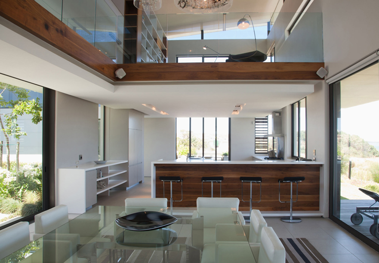 Cocina moderna con barra de madera, comedor con mesa de vidrio y diseño de doble altura con ventanales amplios – FRANCO HomeServices