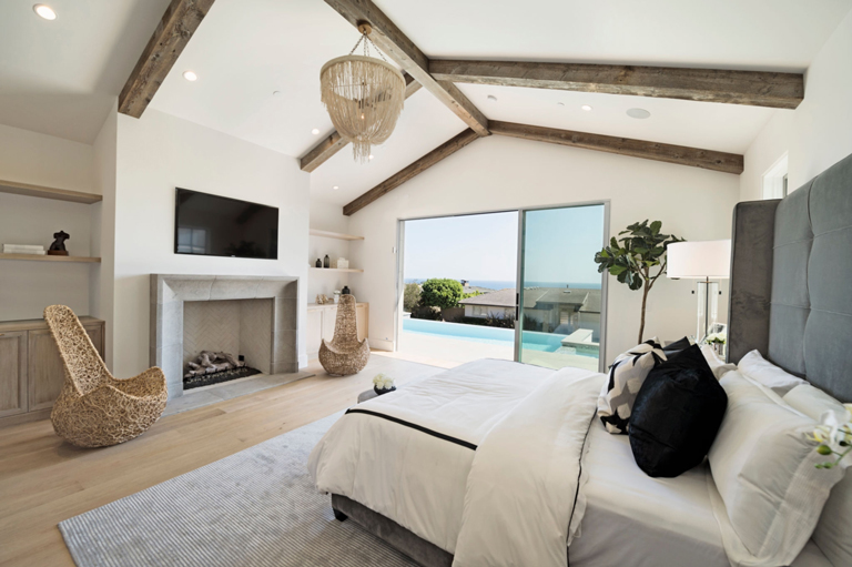 Habitación de lujo con techo de vigas de madera expuestas, chimenea moderna y vista exterior a piscina y mar – FRANCO HomeServices