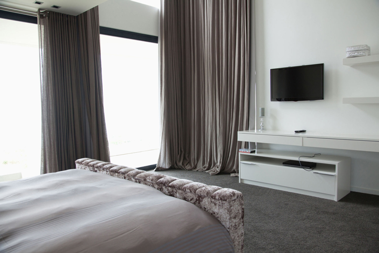Habitación moderna con cortinas de lujo, cama con respaldo tapizado, televisor en pared y mobiliario blanco minimalista – FRANCO HomeServices