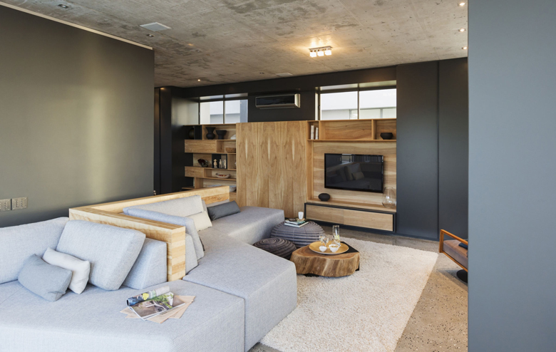 Sala de televisión con muebles de madera natural, sofá modular gris claro y techo en concreto expuesto – FRANCO HomeServices